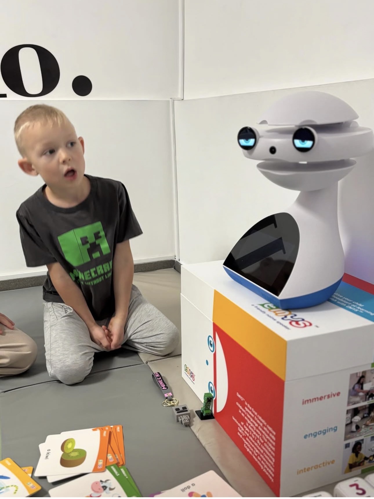 Young boy in Minecraft shirt watching a white robot on a colorful educational box with cards on floor.