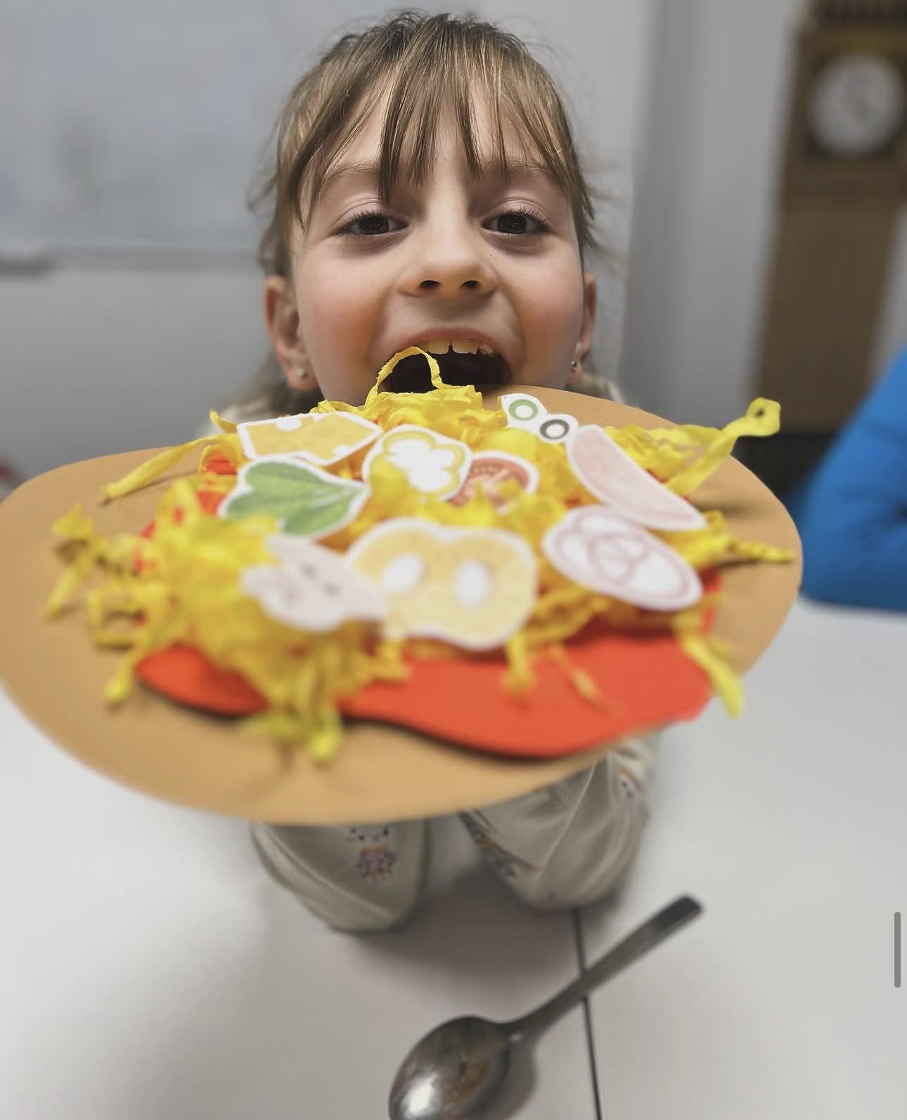 Child holding a colorful craft pizza with paper toppings and yellow shredded paper.
