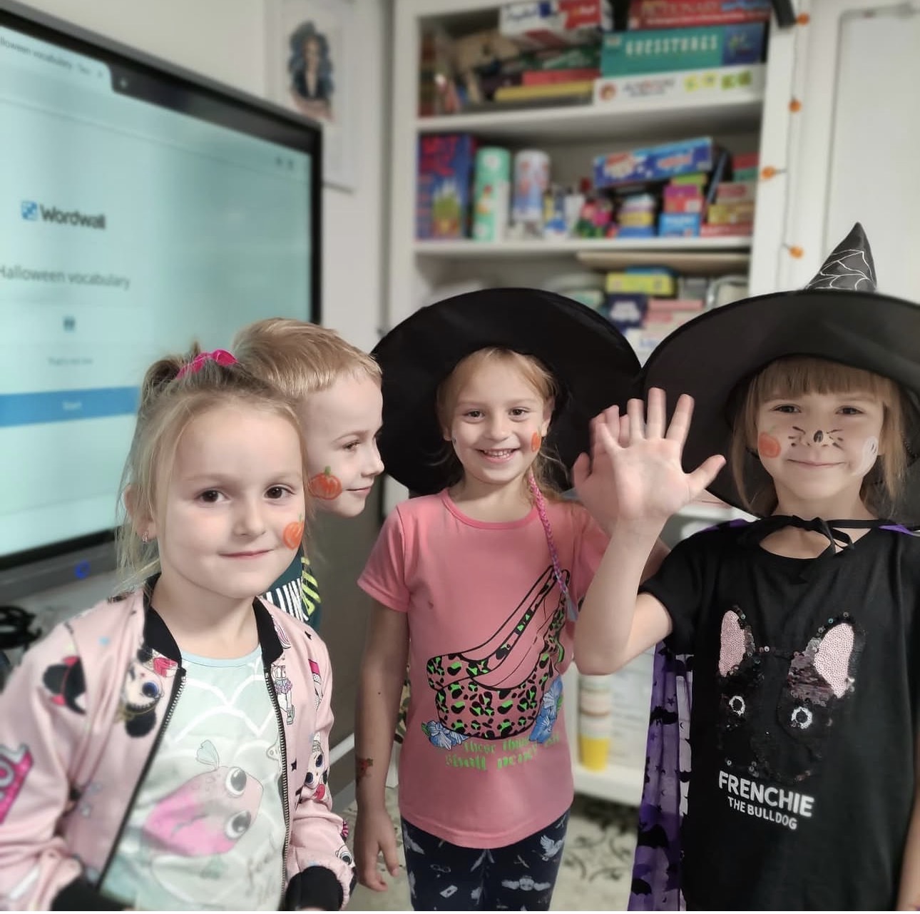 Four smiling children with Halloween face paint and witch hats posing indoors near a screen and shelves.