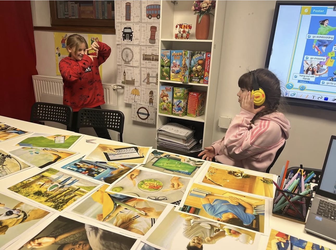 Two girls in a classroom with photos on the table; one wears headphones, the other poses playfully.