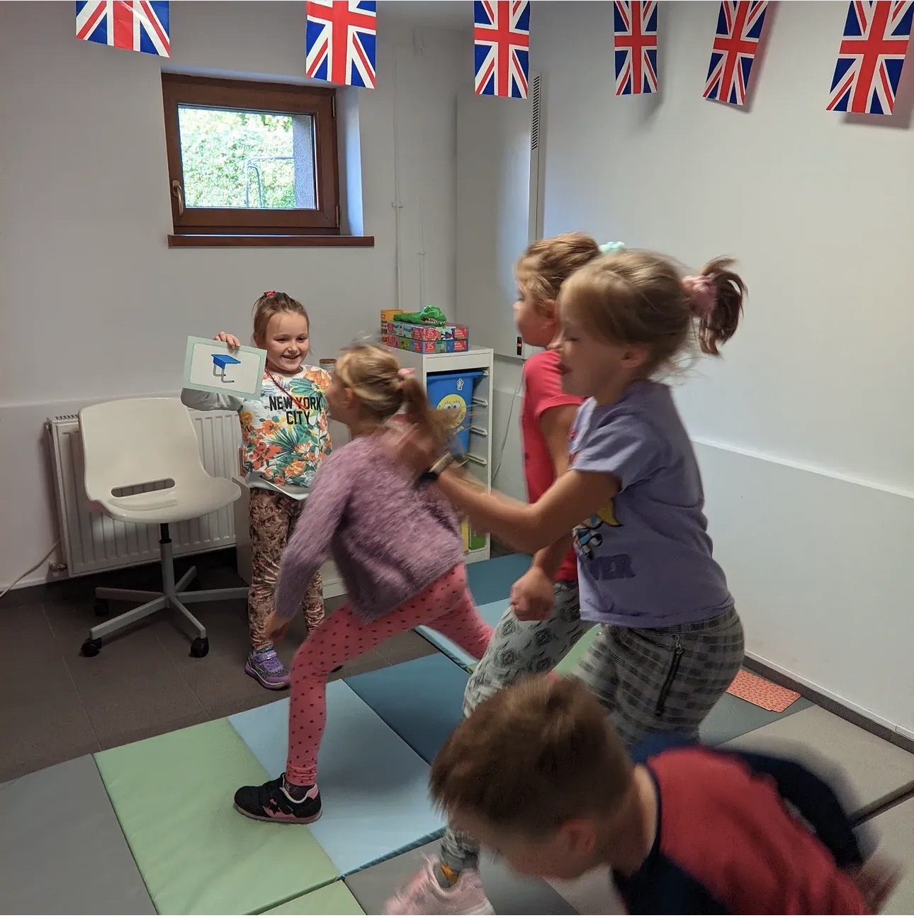 Five children playing indoors with British flags hanging above and one girl holding a picture card.