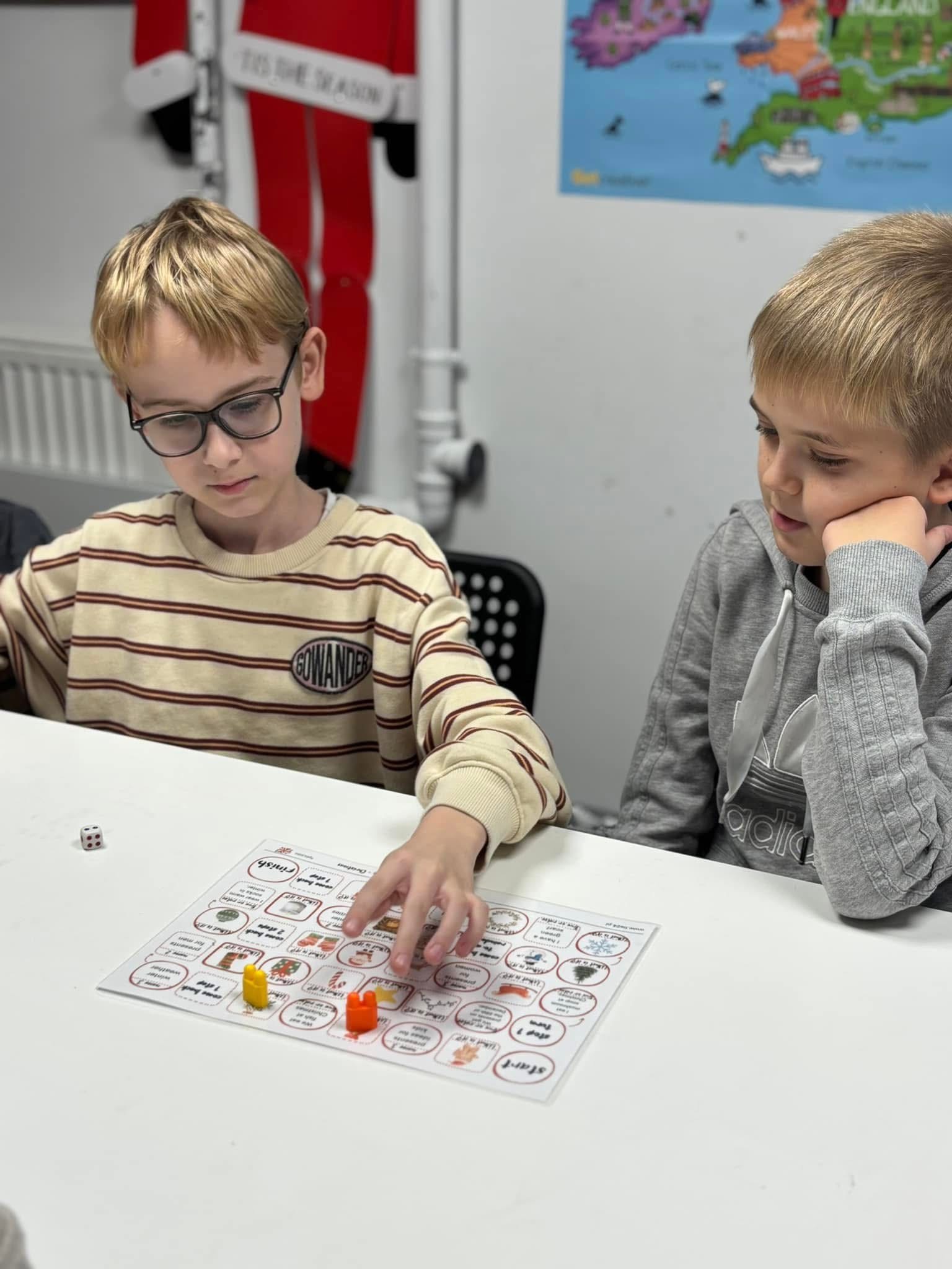 Two boys playing a board game with dice and colorful pieces at a white table indoors.