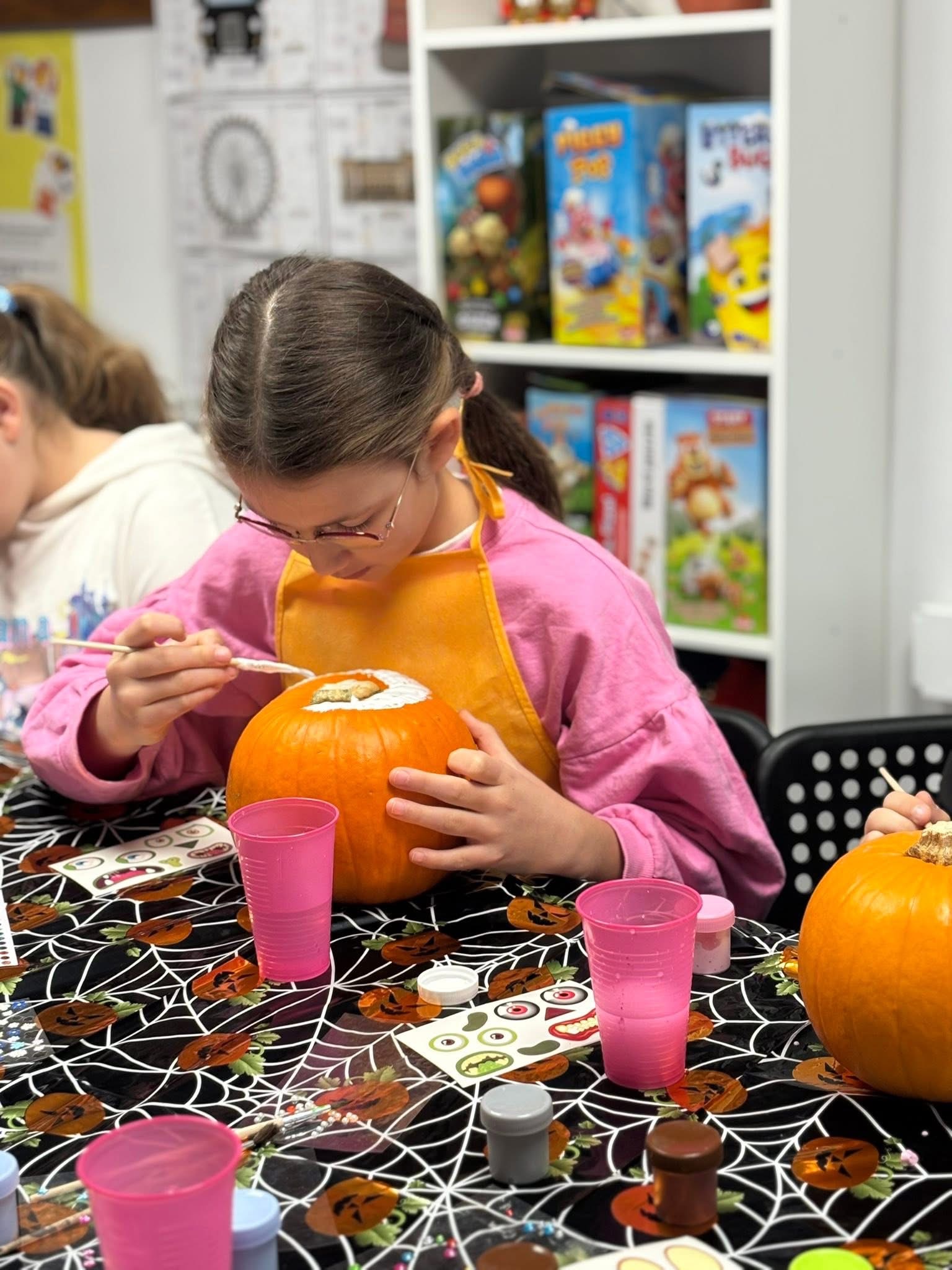Child in glasses and orange apron painting the top of a pumpkin at a decorated table.