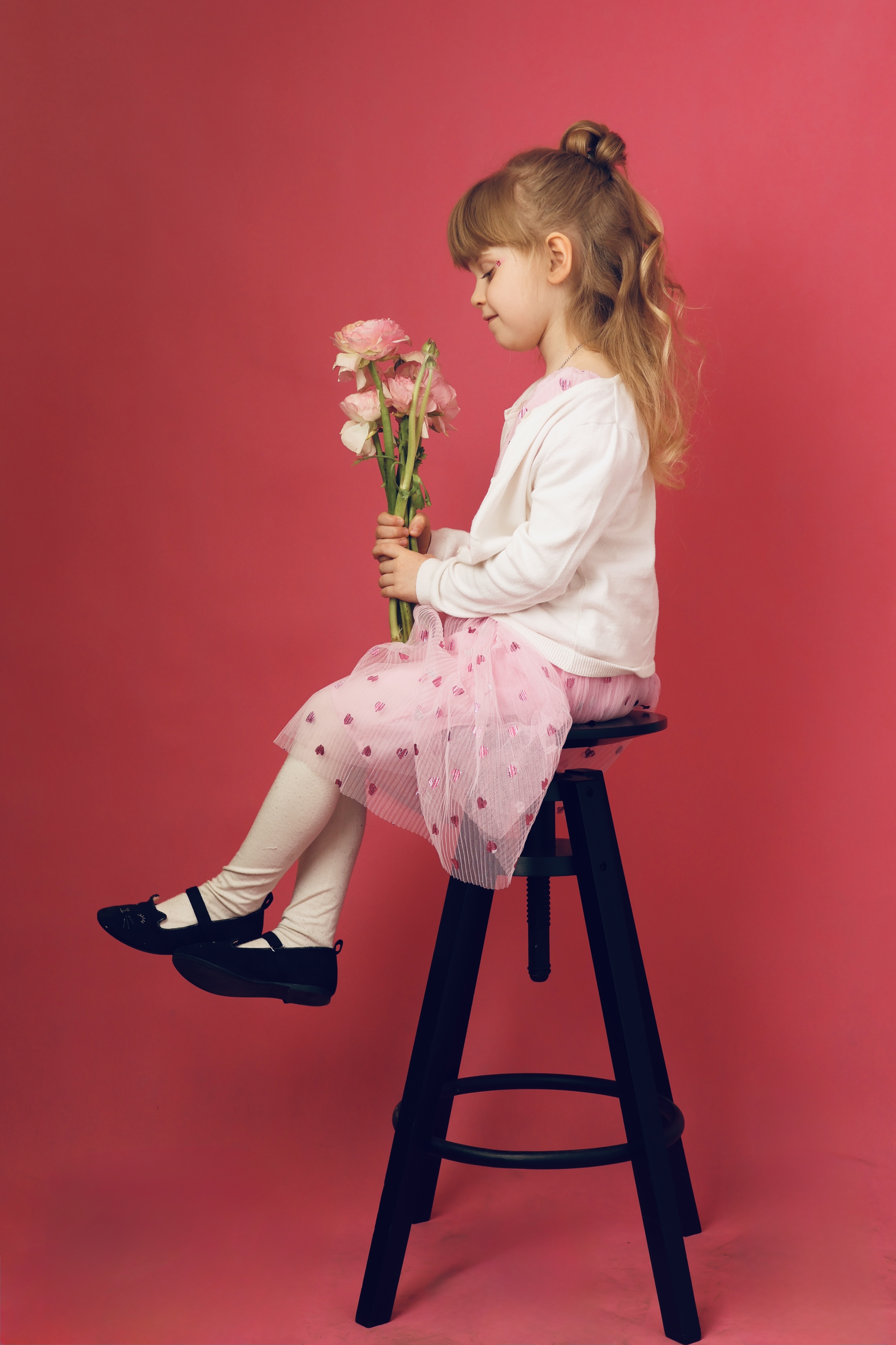 Young girl in pink dress and white cardigan sitting on tall black stool holding pink flowers.