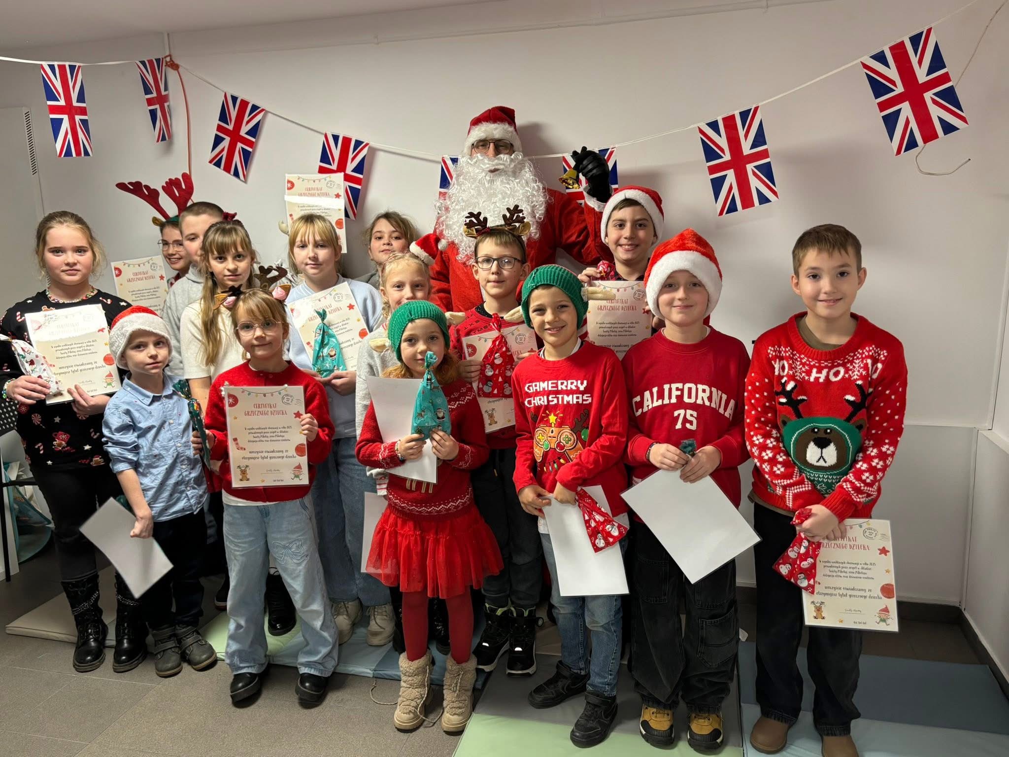 Group of children with Christmas certificates and gifts posing with Santa Claus under British flag bunting.