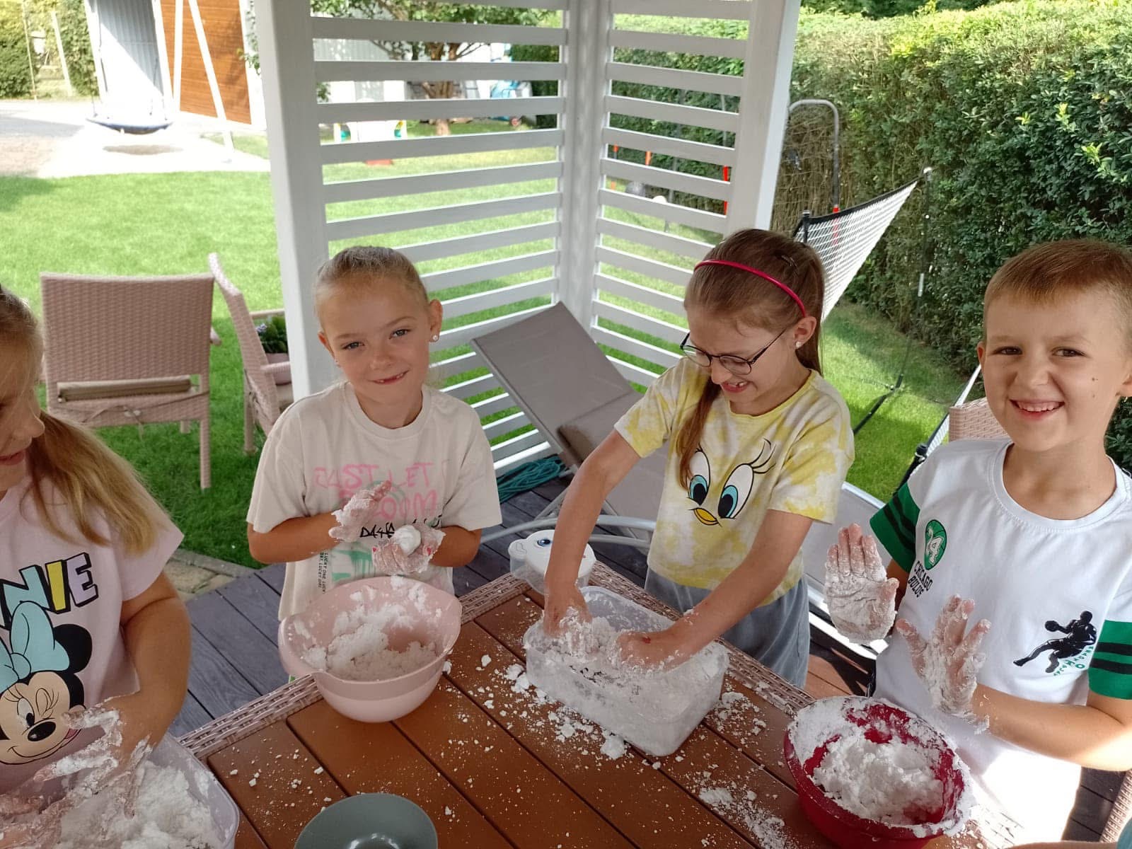 Four children playing with white dough or slime on a wooden table outdoors under a gazebo.