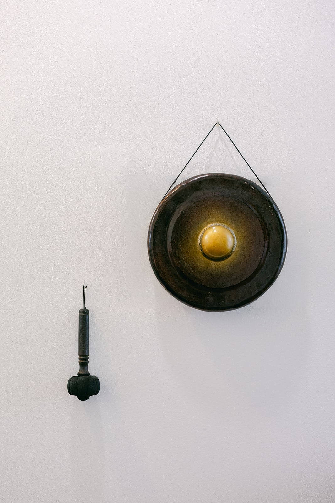 Gong en métal sombre avec un centre doré, suspendu à un mur blanc avec sa mailloche noire à côté.