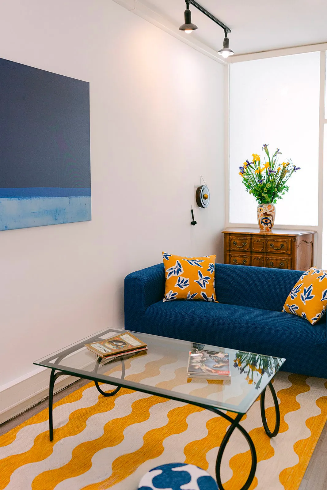 Salon avec canapé bleu, coussins jaunes, table basse en verre et tapis jaune à motifs ondulés.