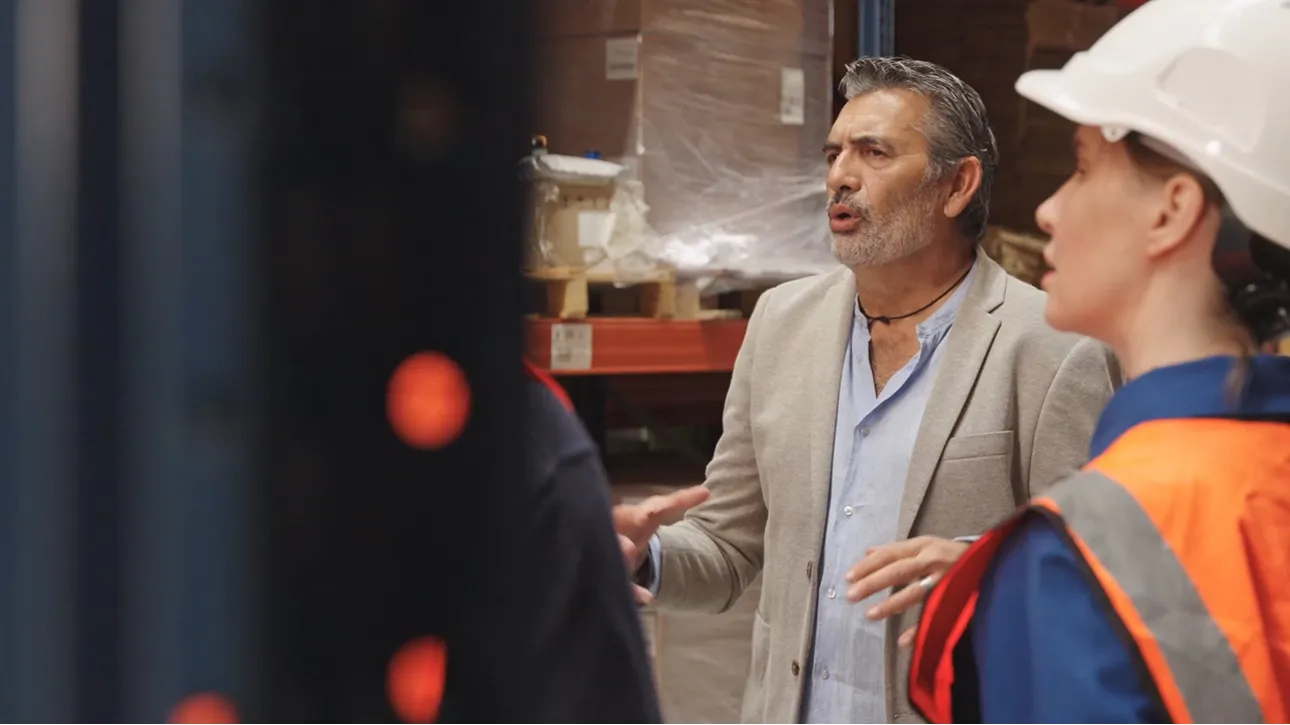 Man in a blazer talking to warehouse workers, one of whom wears a hard hat and safety vest.