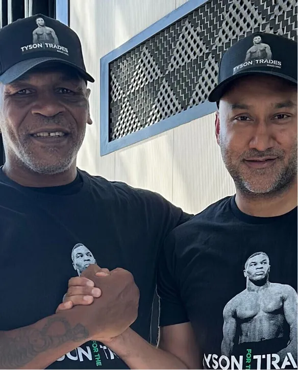 Two men wearing matching Tyson Trades caps and T-shirts shaking hands in front of a textured wall.