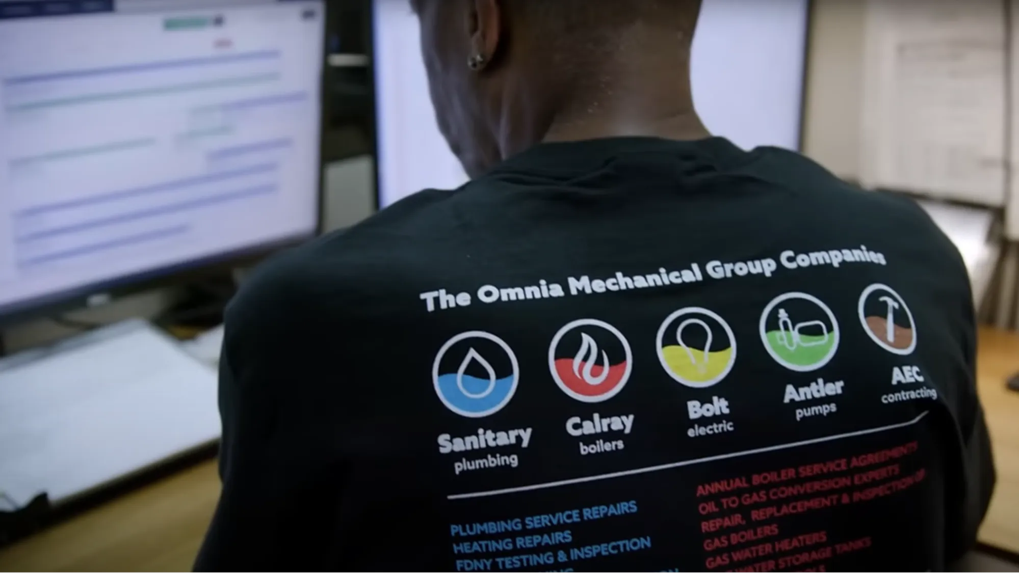 Back view of a person wearing a black shirt featuring logos and services of The Omnia Mechanical Group Companies, including sanitary plumbing, Calray boilers, Bolt electric, Antler pumps, and AEC contracting.