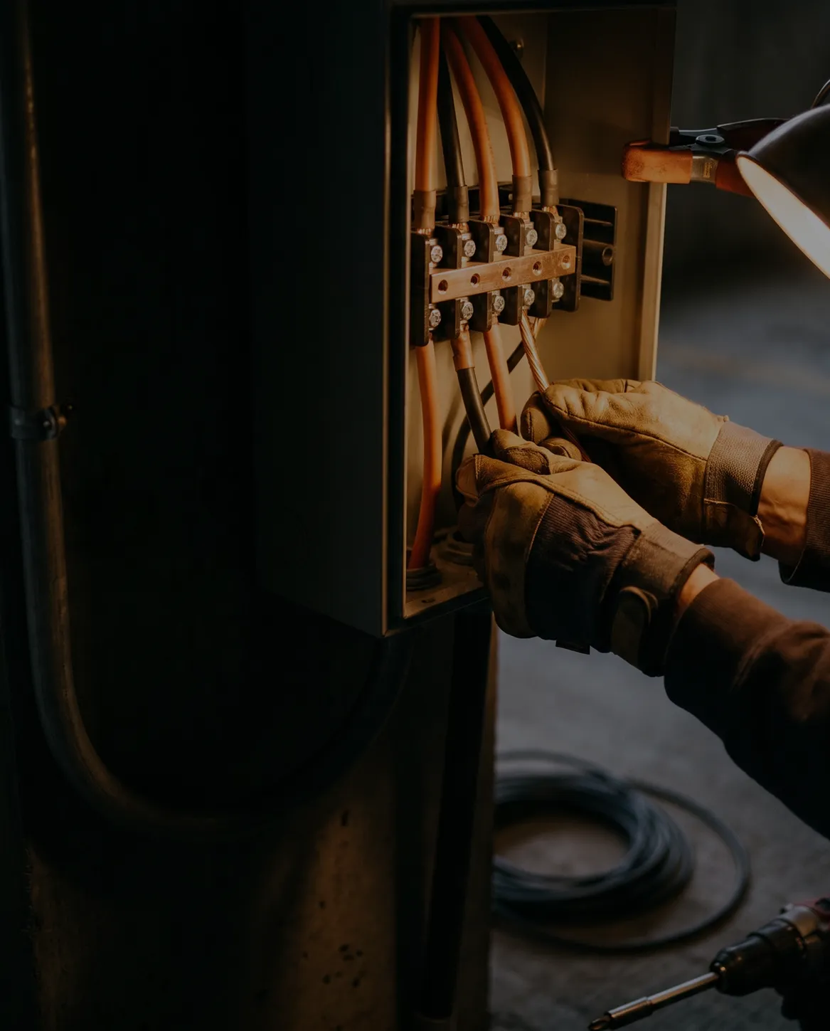 Electrician wearing gloves working on wiring inside an electrical panel with cables and a lamp illuminating the workspace.