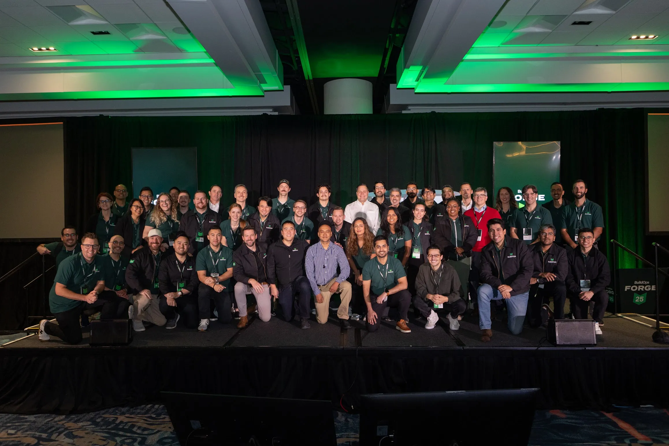 Large group of people posing on stage under green lighting at a BuildOps Forge event.