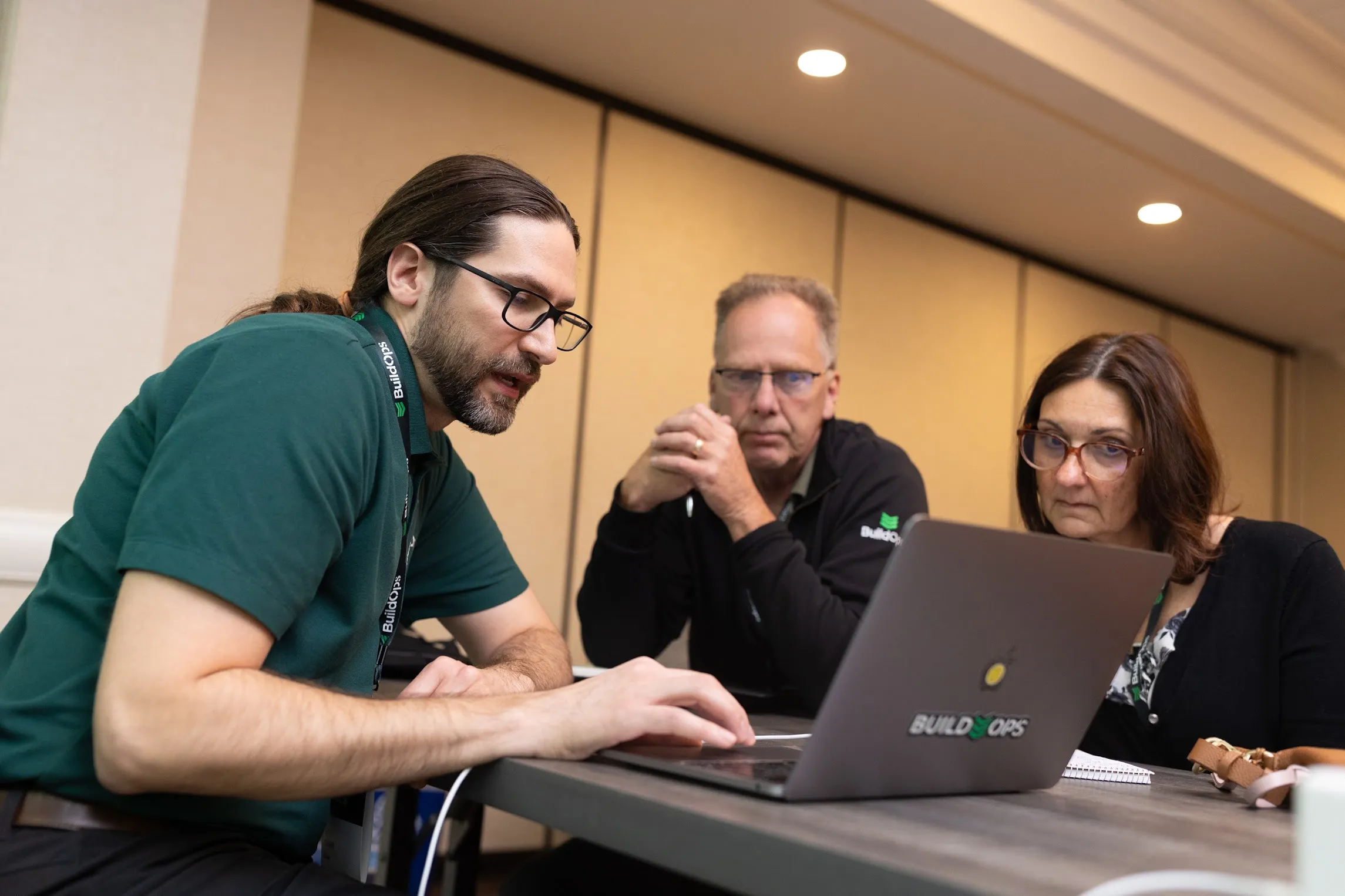 Three adults wearing glasses collaborating at a table, focused on a laptop with BuildOps logo.