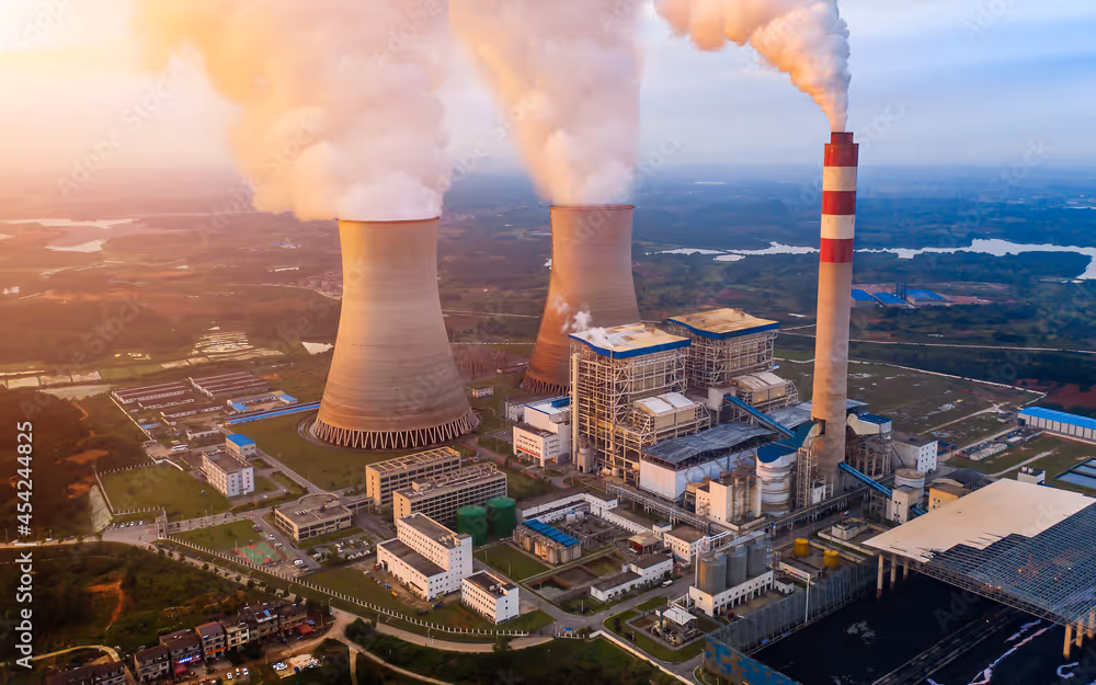 Aerial view of a power plant with cooling towers and a smokestack emitting smoke, surrounded by industrial buildings and green landscape.