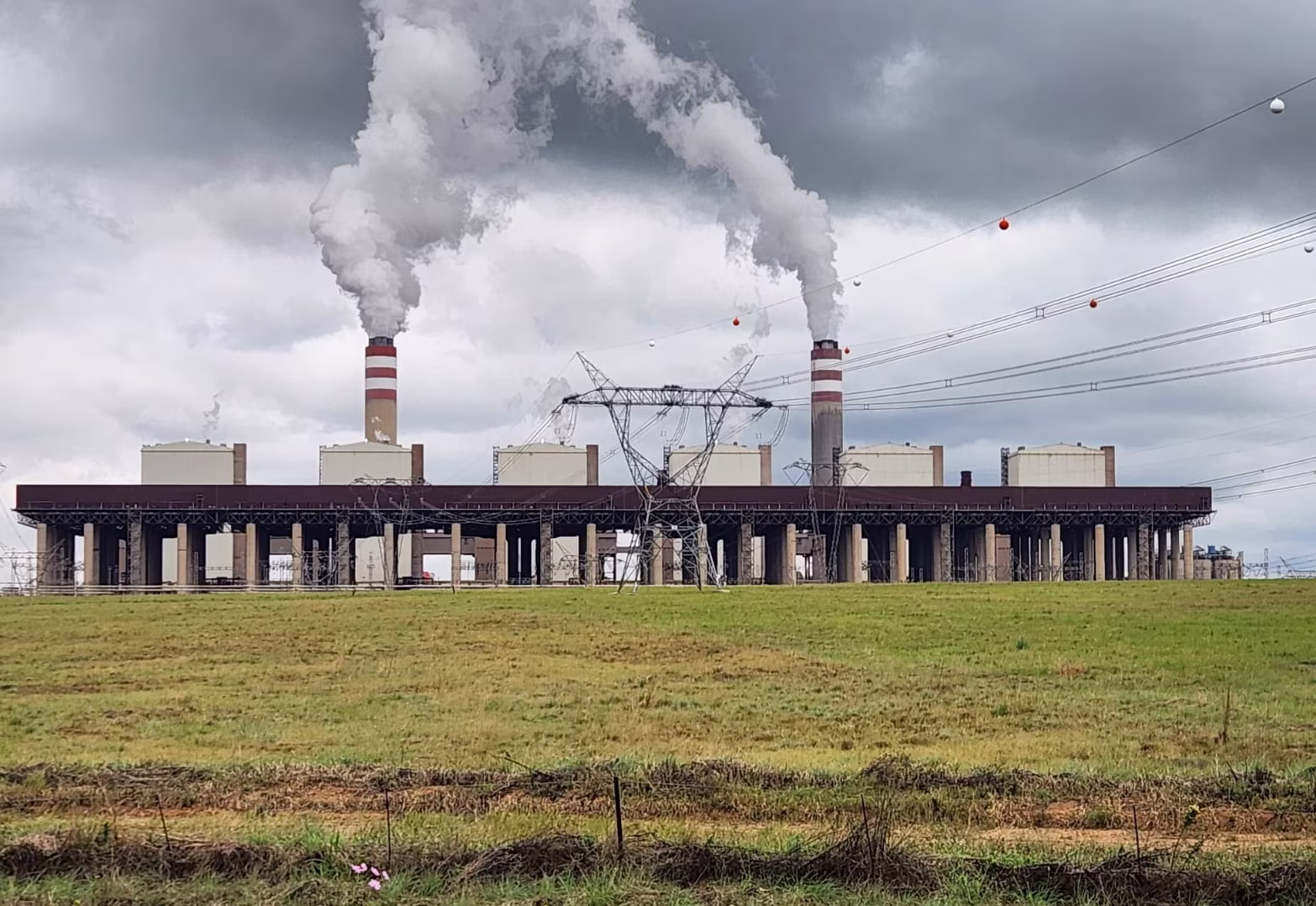 Power plant with two smokestacks emitting smoke against a cloudy sky over a grassy field.