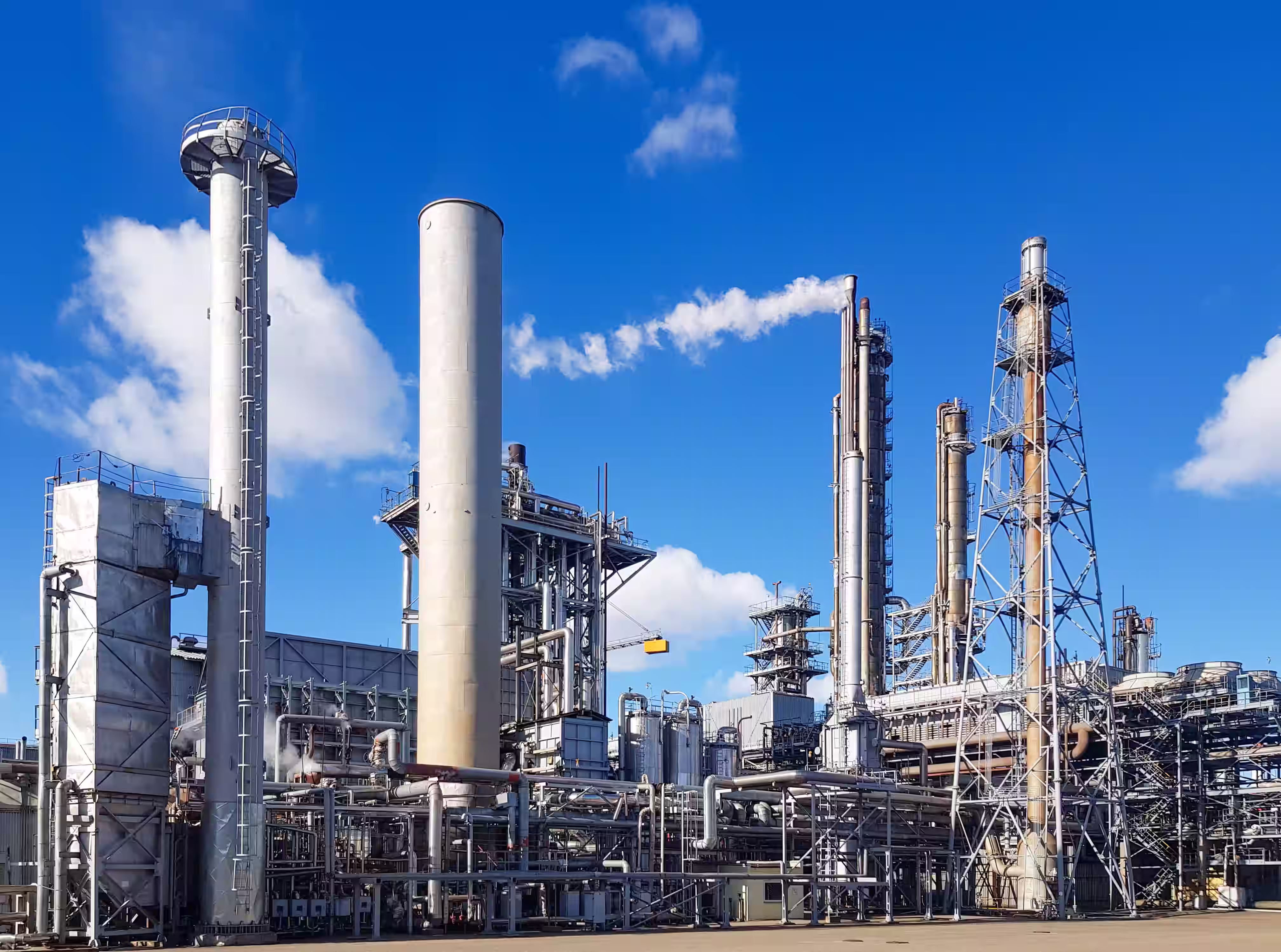 Large industrial refinery with metal towers and pipes under a blue sky with white clouds.