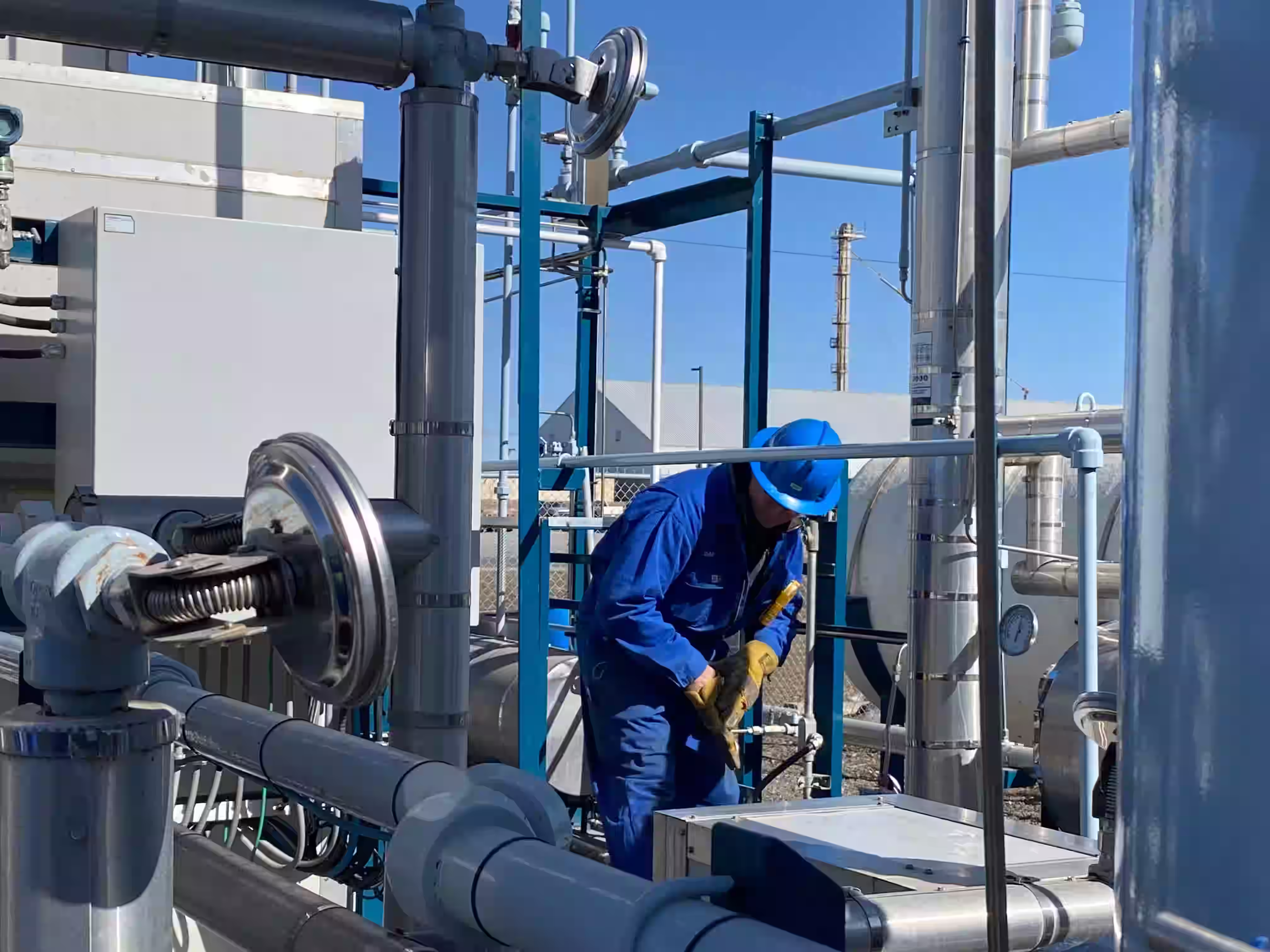 Industrial worker in blue coveralls and hard hat using a wrench to work on metal pipes at an outdoor facility under clear sky.