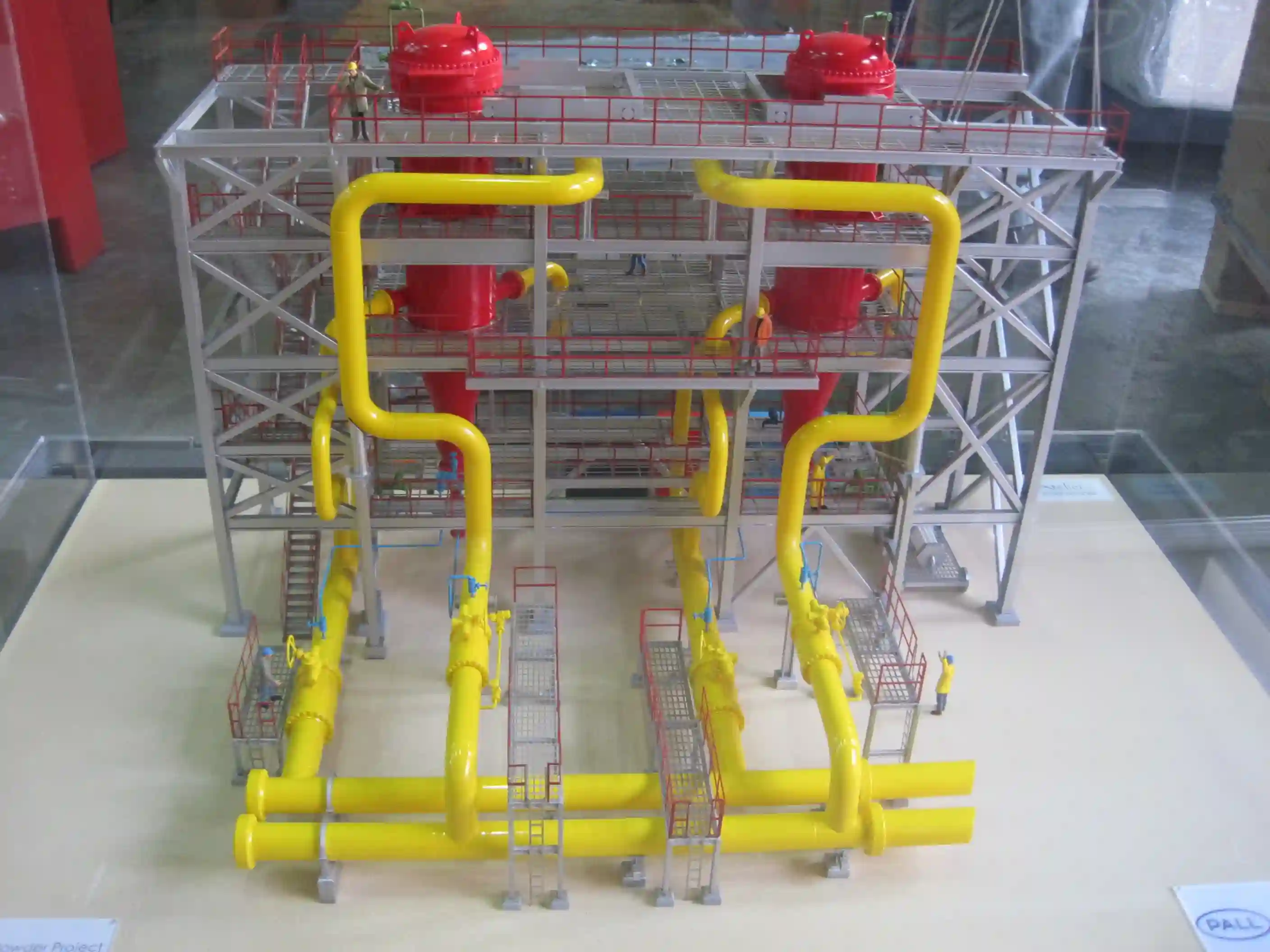 Scale model of an industrial filtration system with yellow pipes, red cylindrical filters, and metal framework with miniature worker figures.