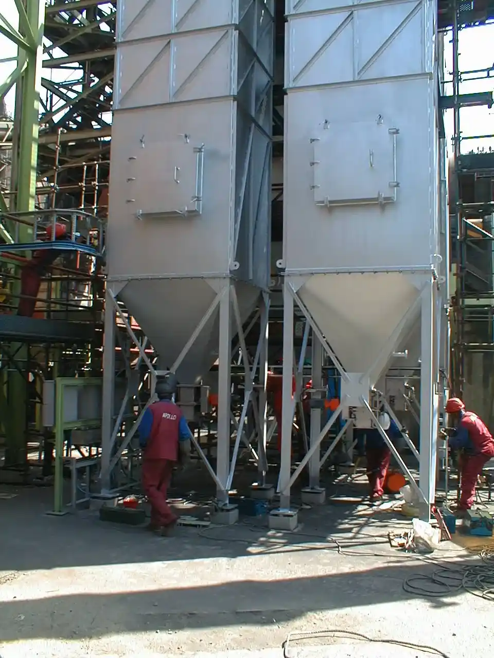 Industrial scene with workers in red uniforms around large gray metal silos in a factory setting.