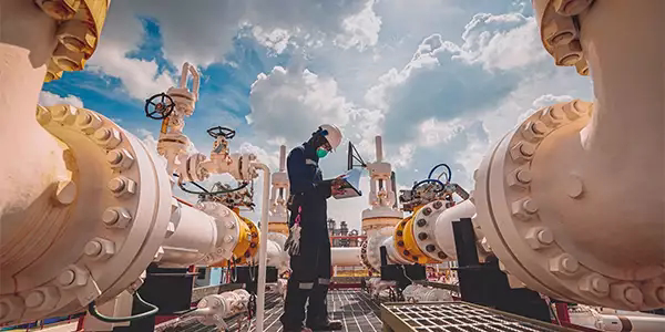 Worker in protective gear inspecting large industrial oil and gas pipelines under a blue sky with clouds.