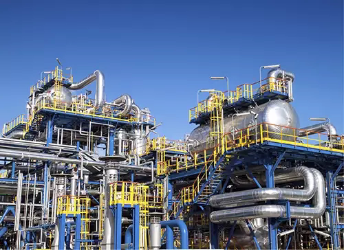Industrial chemical processing plant with large metal tanks, pipes, and yellow railings under a clear blue sky.