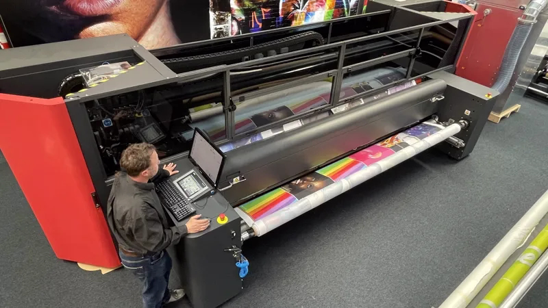 Operator monitoring a large industrial printer producing colorful printed material on wide rolls of paper.