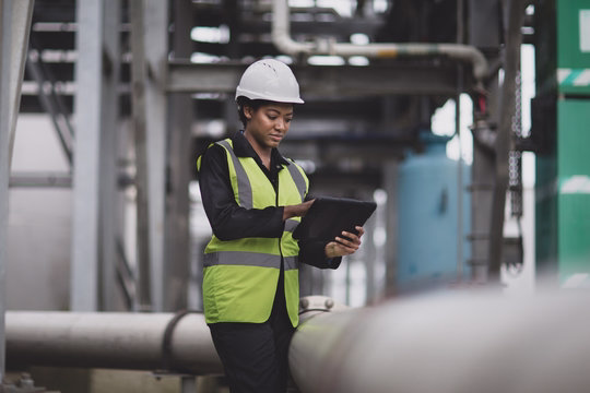 Female industrial worker wearing a white hard hat and reflective vest using a tablet in a factory setting.