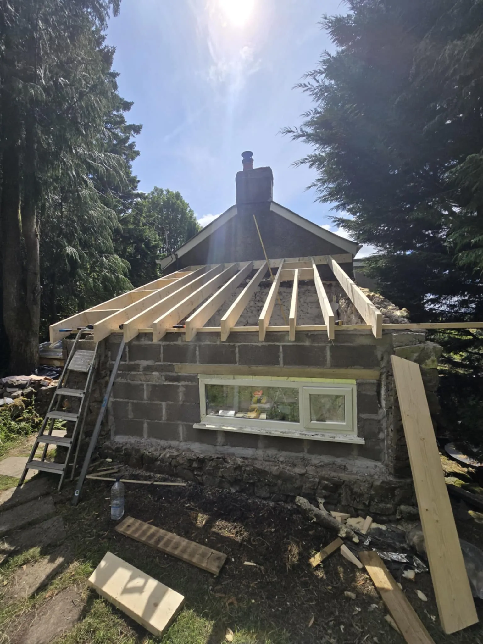 House under construction with wooden beams framing a new roof above a stone wall and a narrow horizontal window.