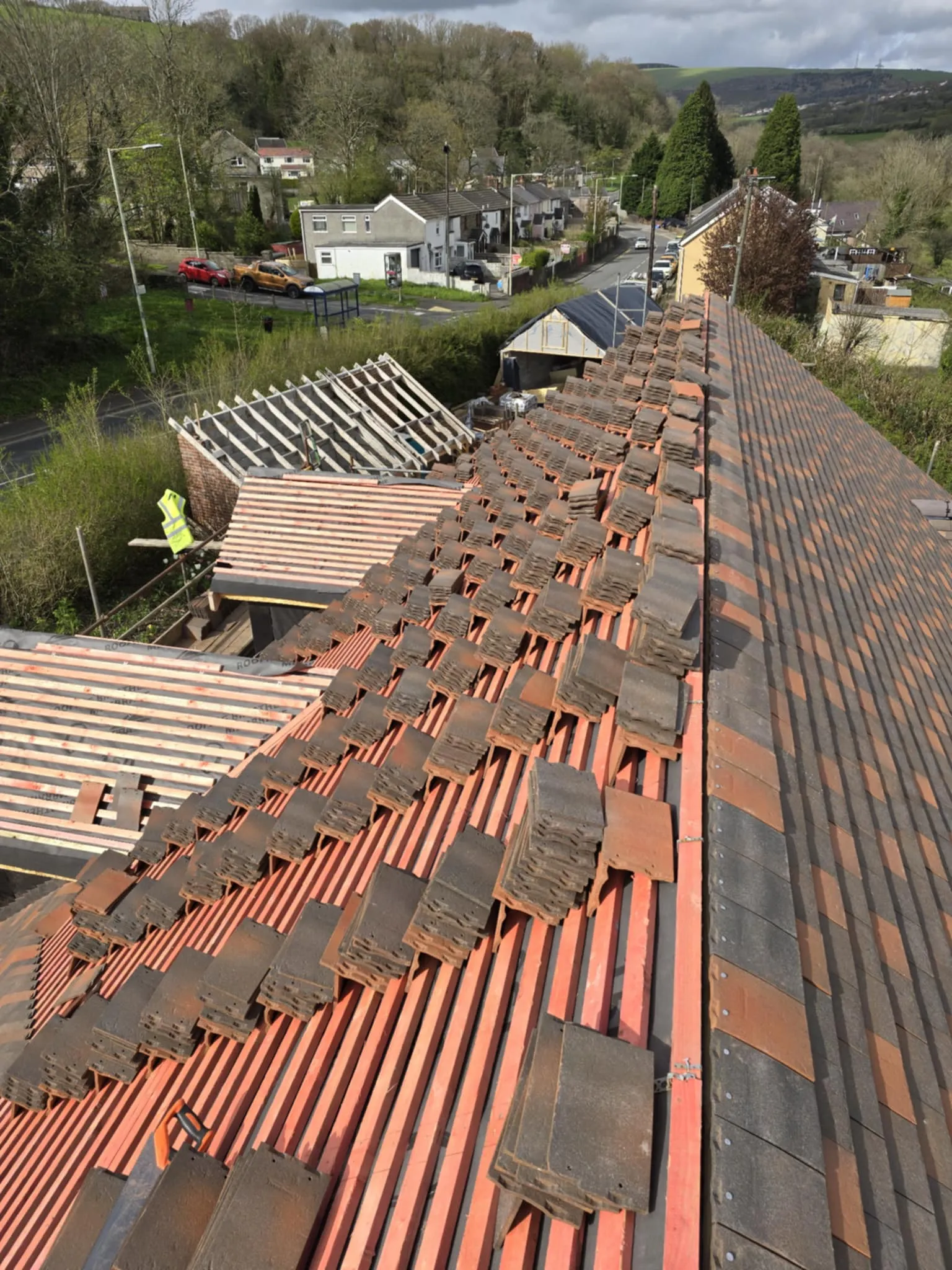 Rooftop under construction with stacks of roof tiles placed on wooden beams, overlooking a suburban neighborhood and wooded hills.