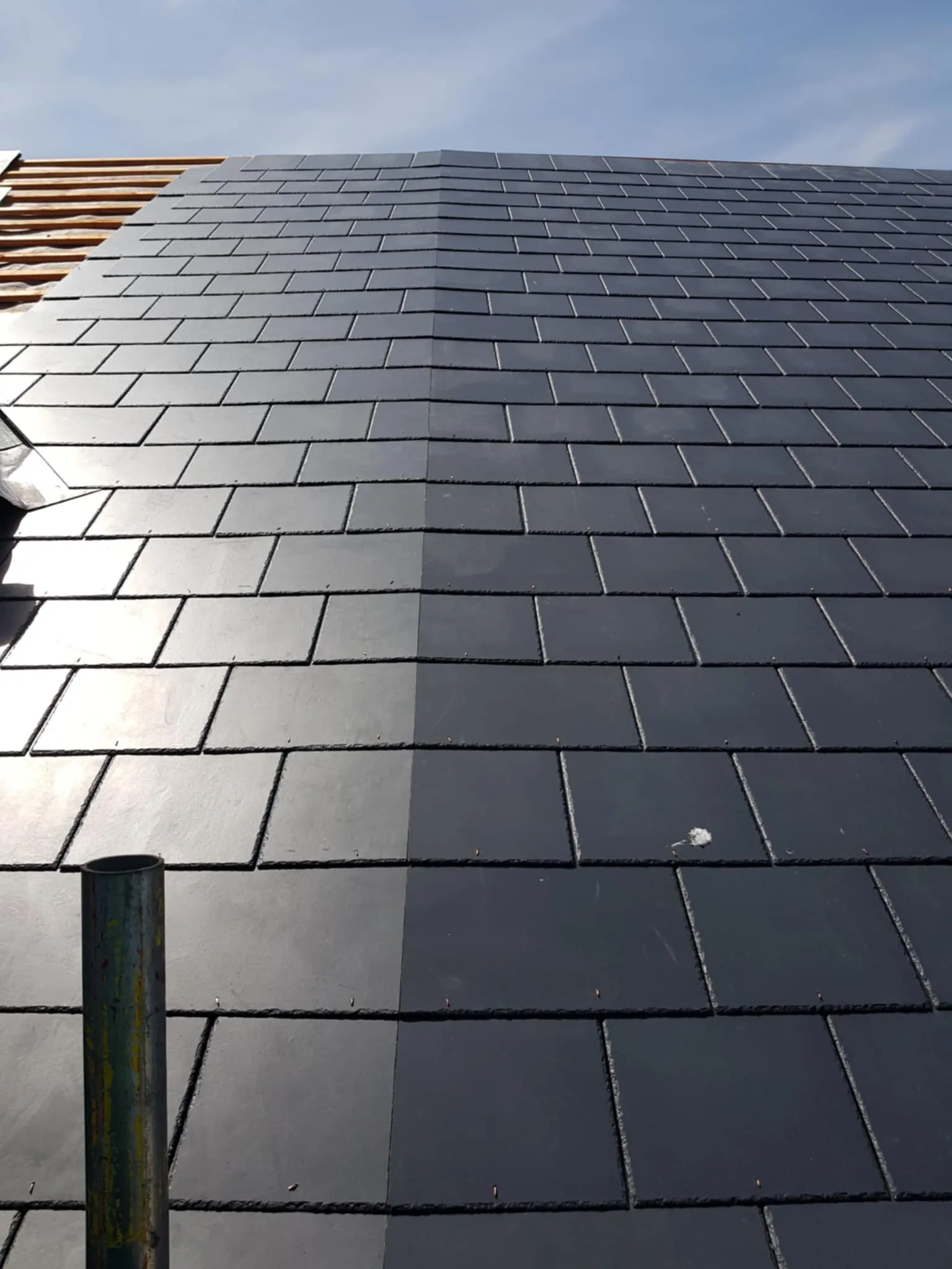Close-up of a roof with newly installed dark gray rectangular tiles with a reflective surface under a blue sky.