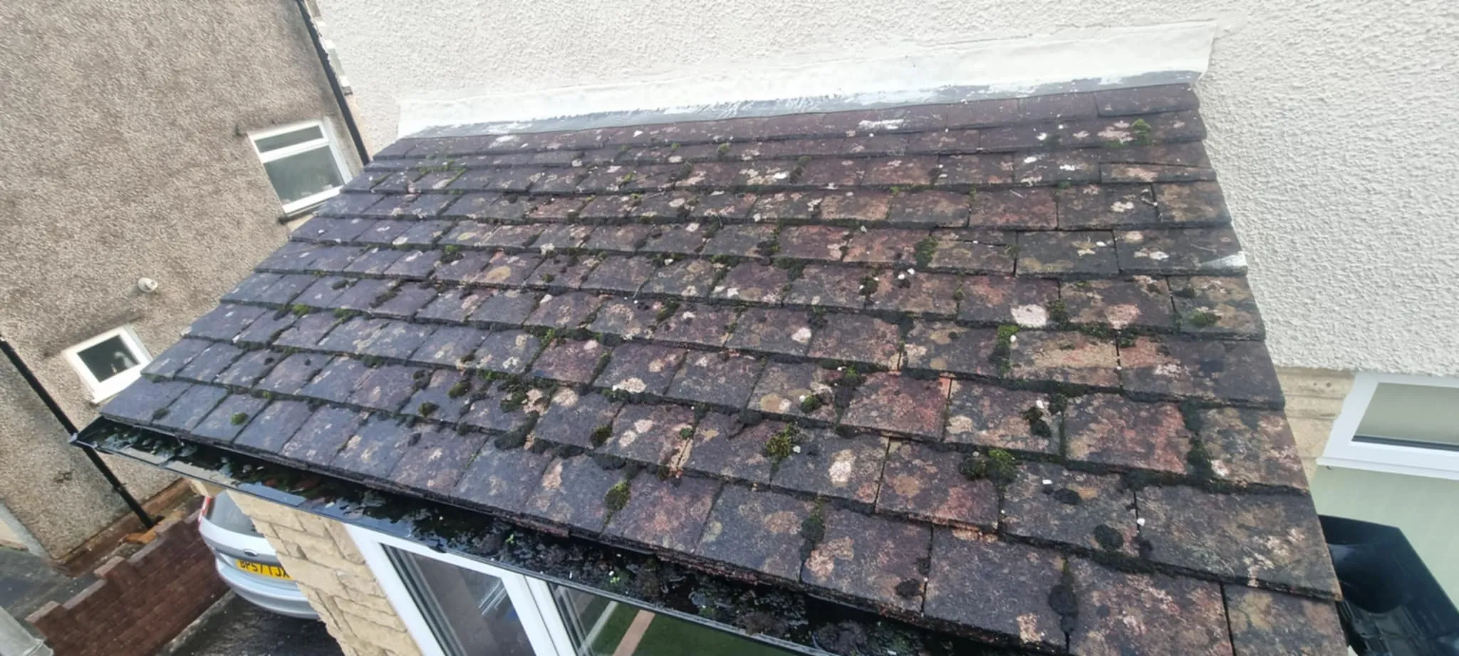 Close-up of an aged tiled roof with moss and lichen patches, attached to a building with beige textured walls and nearby windows.