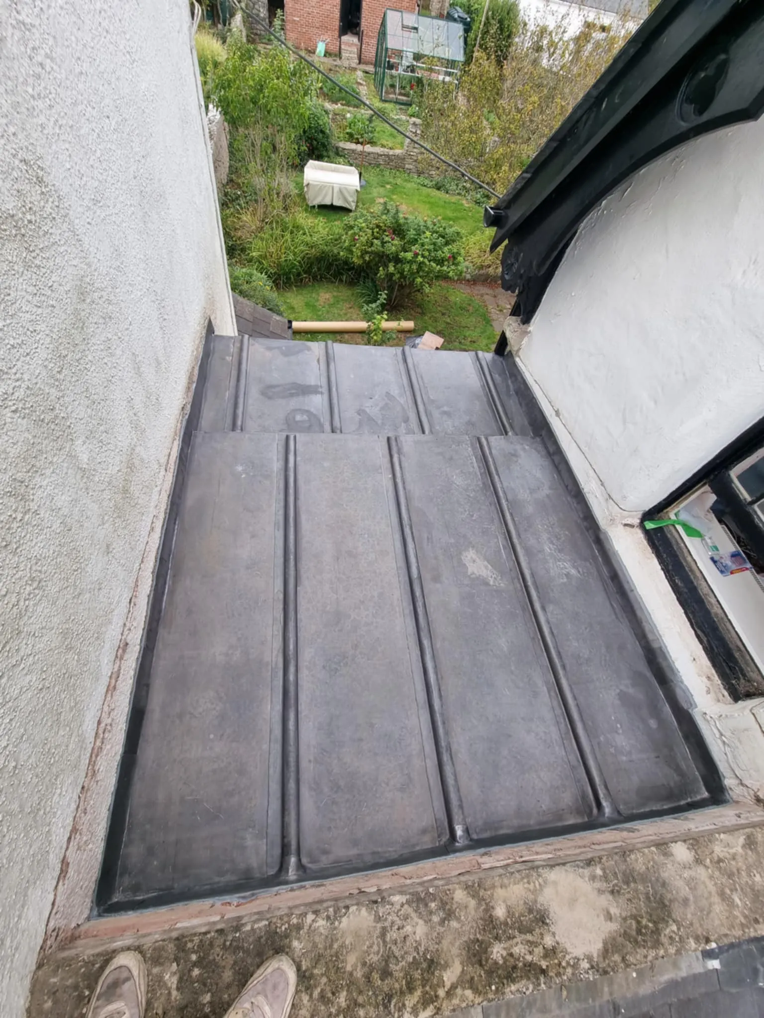 View looking down at a metal roof panel installation on a residential building with a garden in the background.