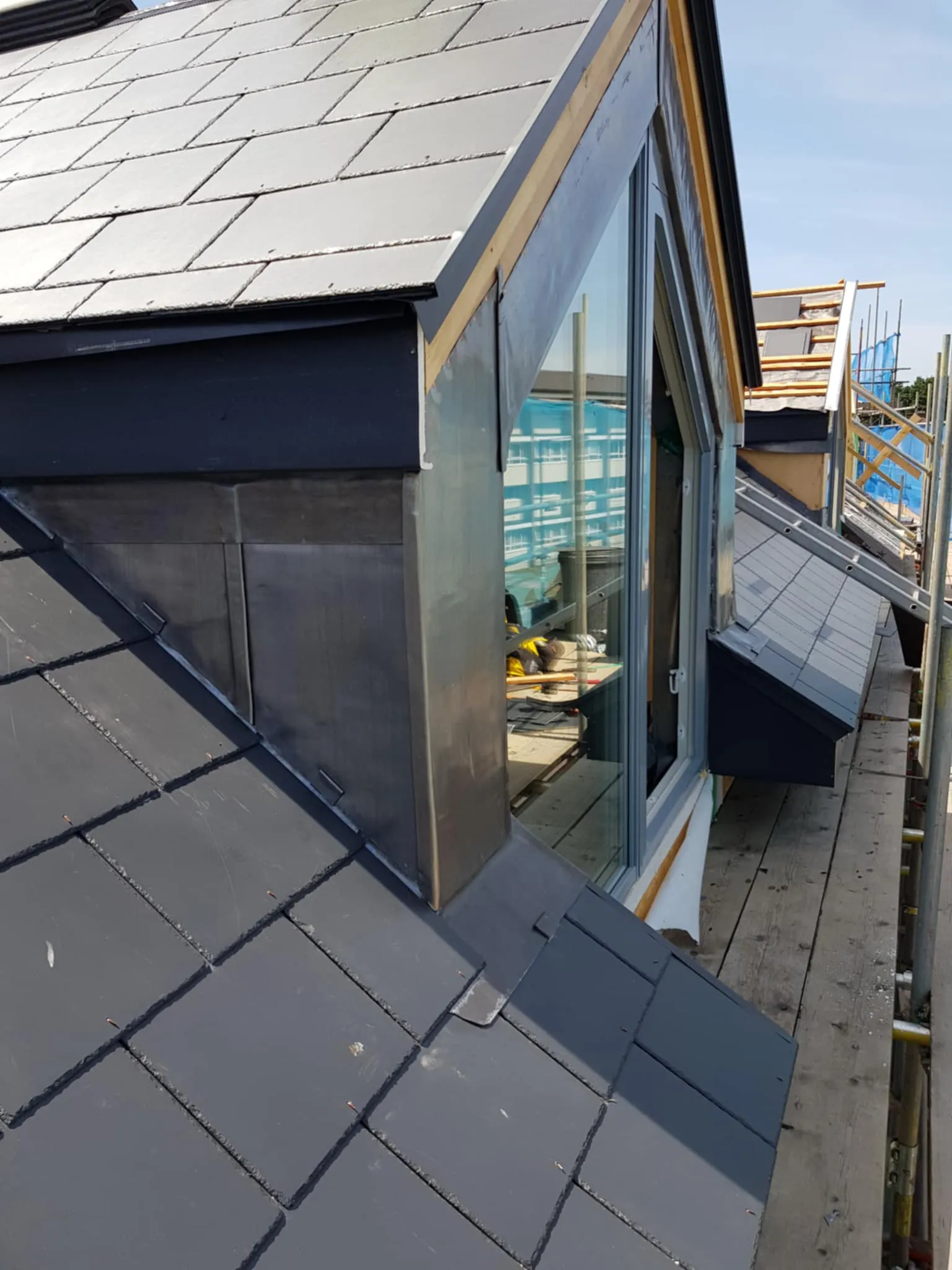Close-up of a pitched roof with dark gray tiles and two dormer windows under construction, with scaffolding and wooden planks.