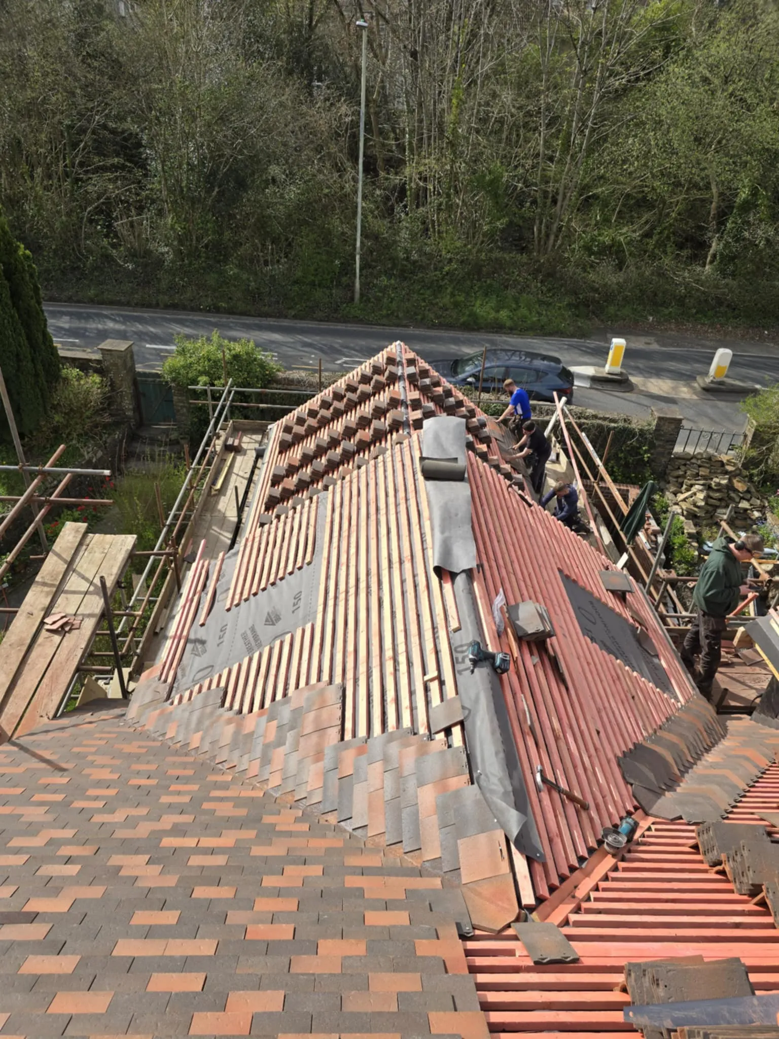 View from above of a roof under construction with workers installing red tiles on wooden battens surrounded by scaffolding and trees.