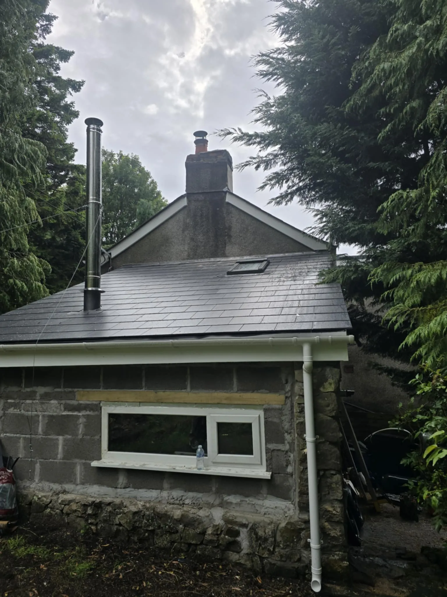 Small stone and concrete building with a dark slate roof, chimney, a skylight, and surrounded by tall green trees.