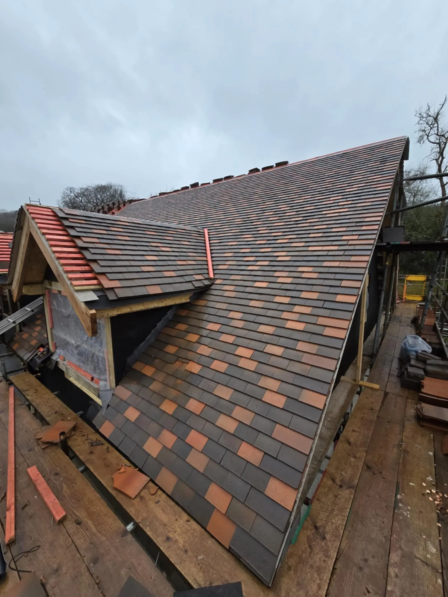Newly installed roof with dark gray and brownish-red tiles on a cloudy day, viewed from scaffolding.