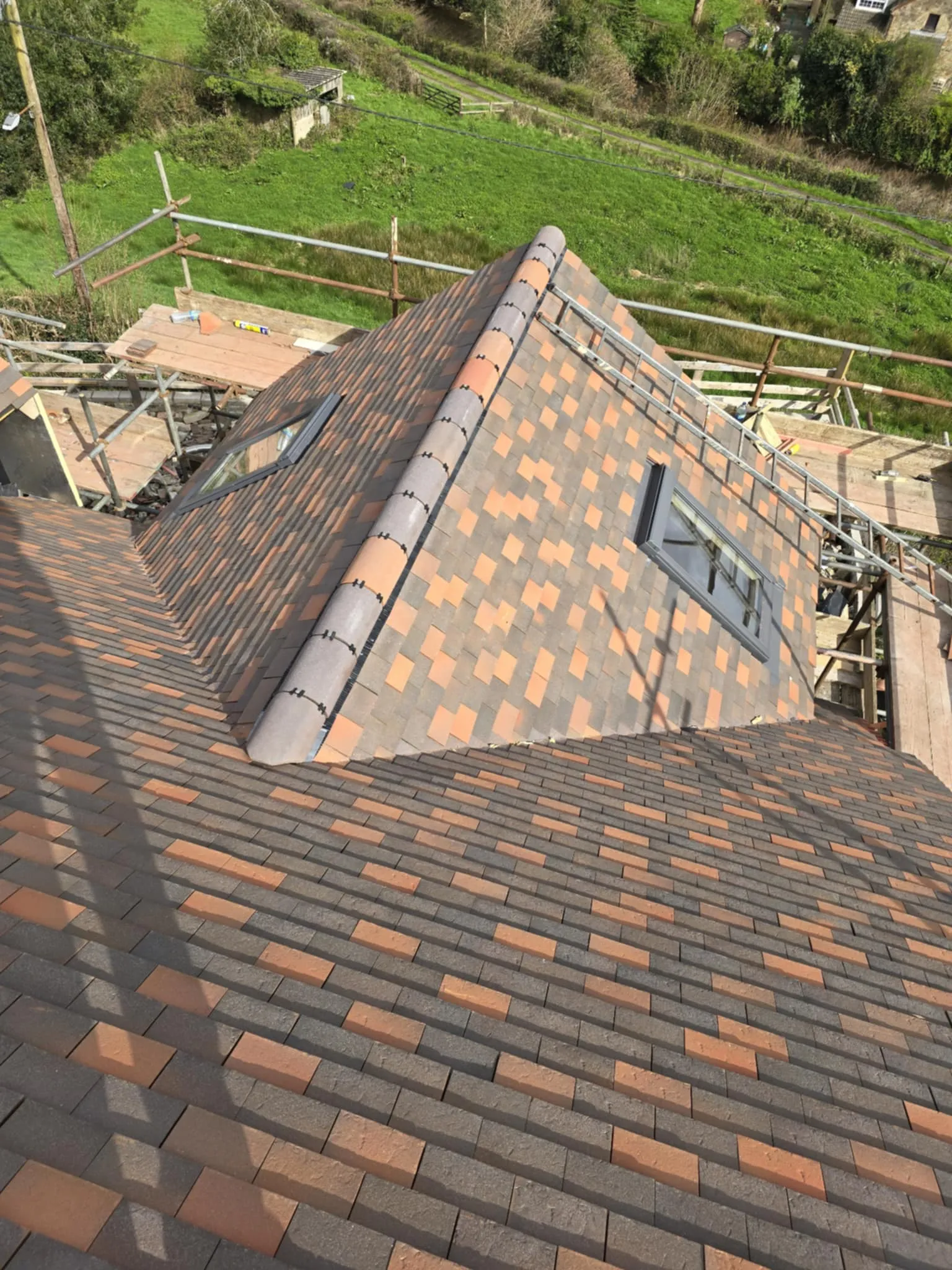 Newly installed residential roof with orange and gray tiles and two skylights surrounded by scaffolding and green fields.