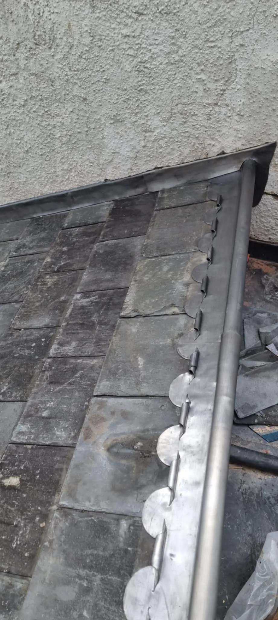 Close-up of a roof edge with overlapping dark shingles next to a silver metal gutter with circular tabs along the side.