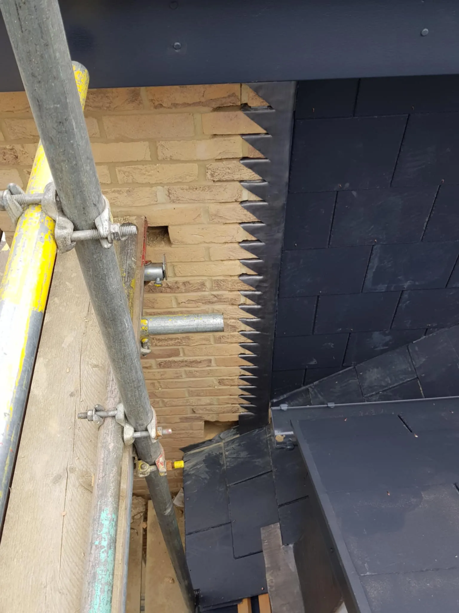 Close-up of black roof tiles meeting a brick wall with metal flashing in a sawtooth pattern, viewed from scaffolding.