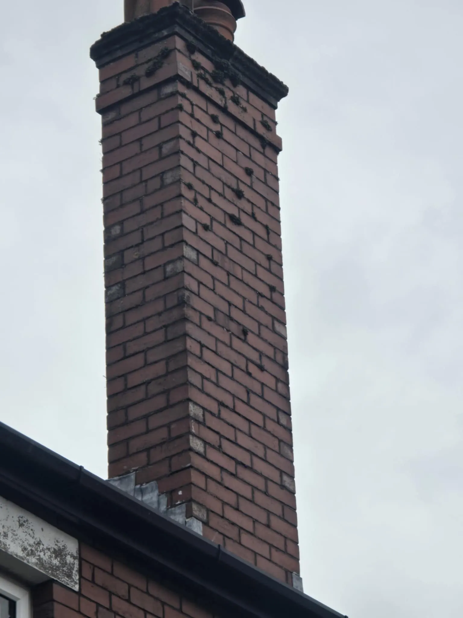 Tall red brick chimney with some moss growth extending from a house roof against a cloudy sky.
