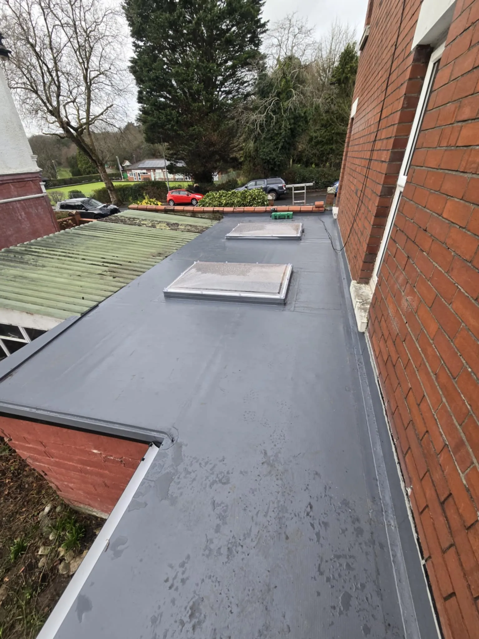 Flat gray roof with two rectangular skylights attached to a red brick building with a green corrugated roof adjacent.