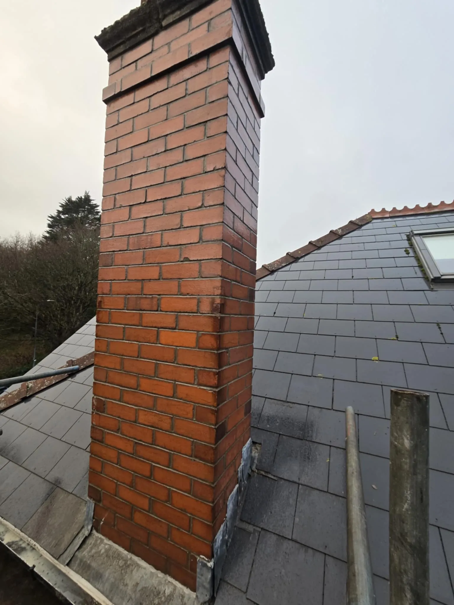 Tall red brick chimney on a gray tiled sloped roof with a small skylight window and some metal scaffolding poles.