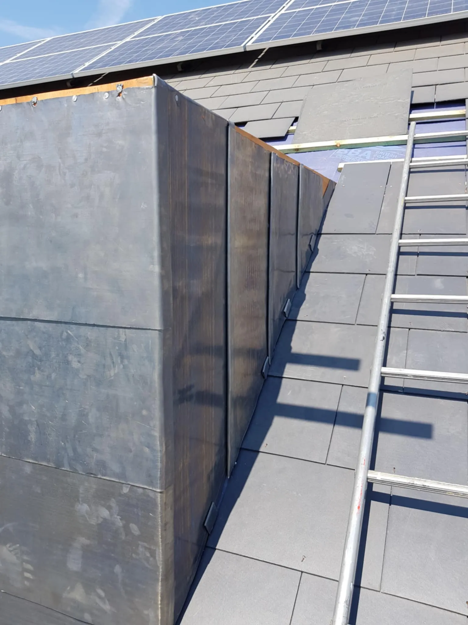 Roof with solar panels and a metal chimney covering next to a ladder on gray slate tiles under clear blue sky.