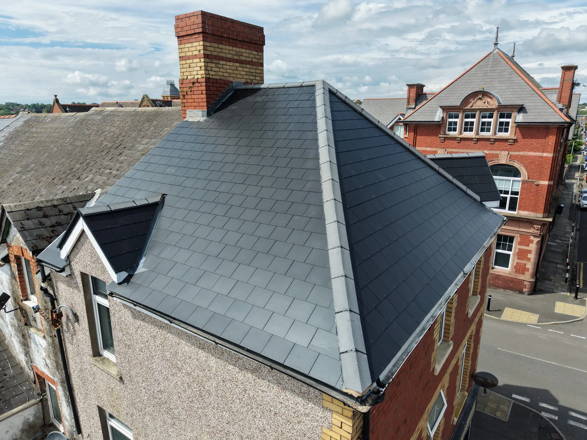 Aerial view of a building with a dark gray slate roof and a brick chimney, surrounded by adjacent buildings on a city street.