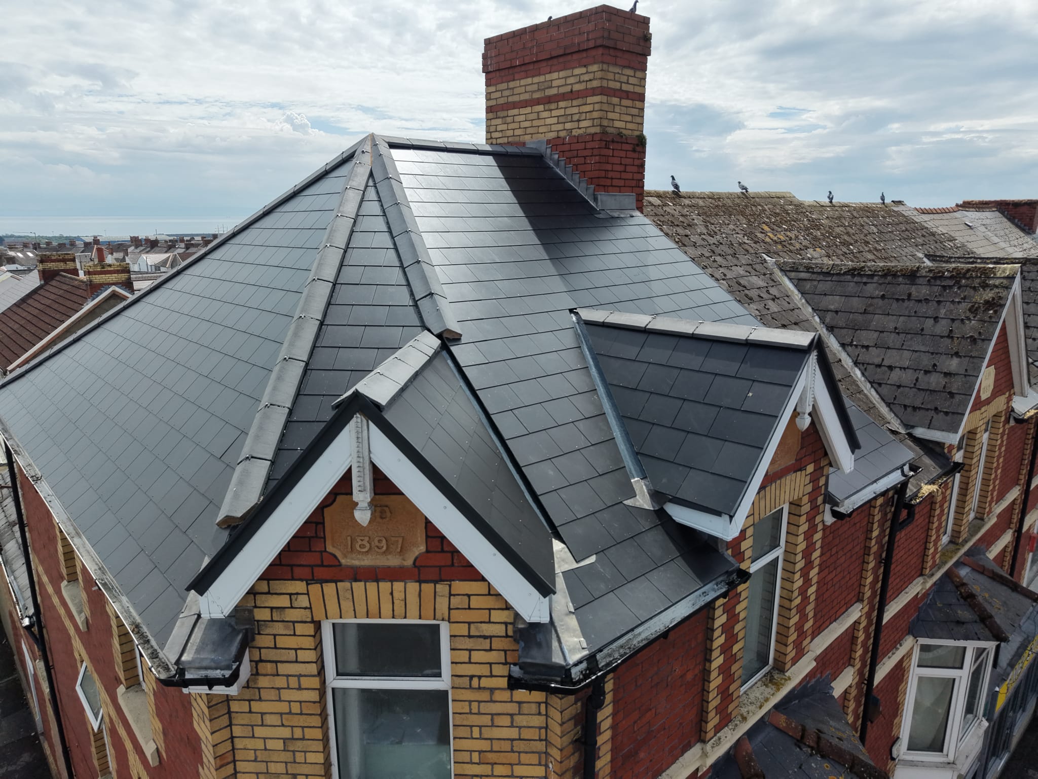 Corner of a brick house with a newly installed dark grey slate roof and a red brick chimney, with other older rooftops visible in the background.