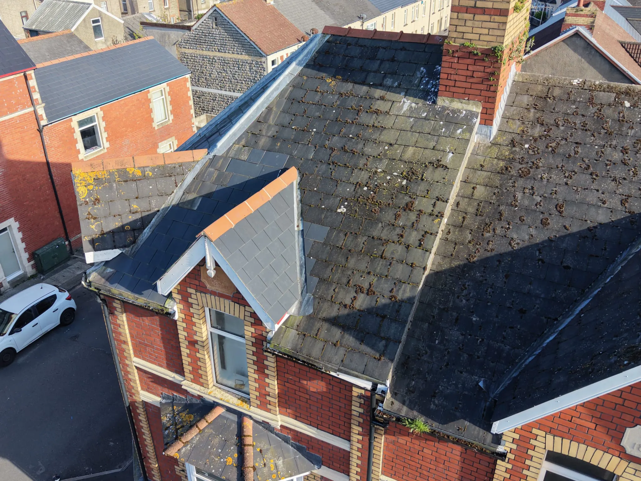 Aerial view of slate-tiled rooftops on brick residential buildings with moss and lichen patches, adjacent to a street with a white car.
