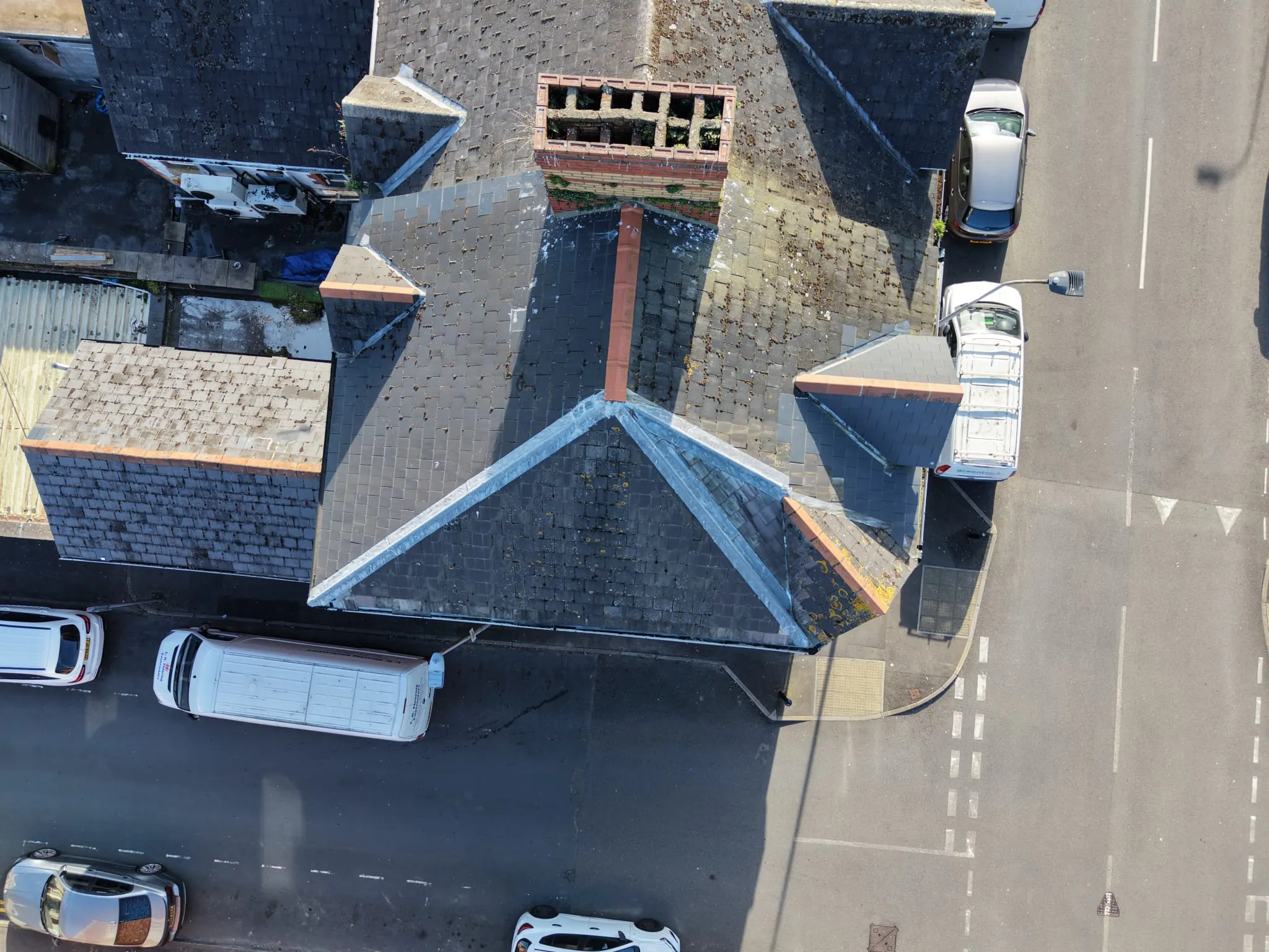 Aerial view of a building rooftop with multiple pitched roofs, chimneys, and several parked cars on adjacent streets.