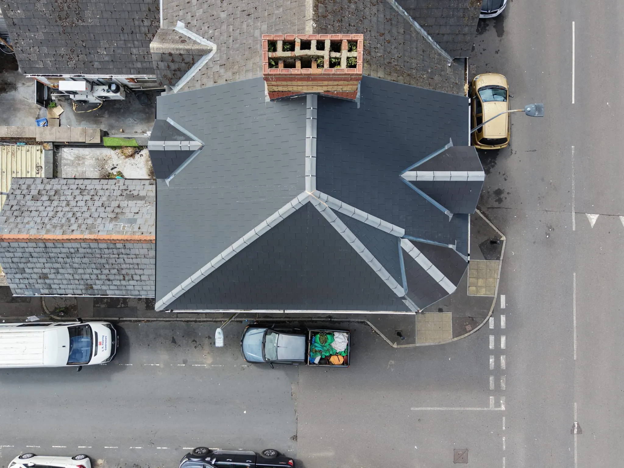 Aerial view of a building with a dark slate roof, surrounded by parked vehicles on adjacent streets.