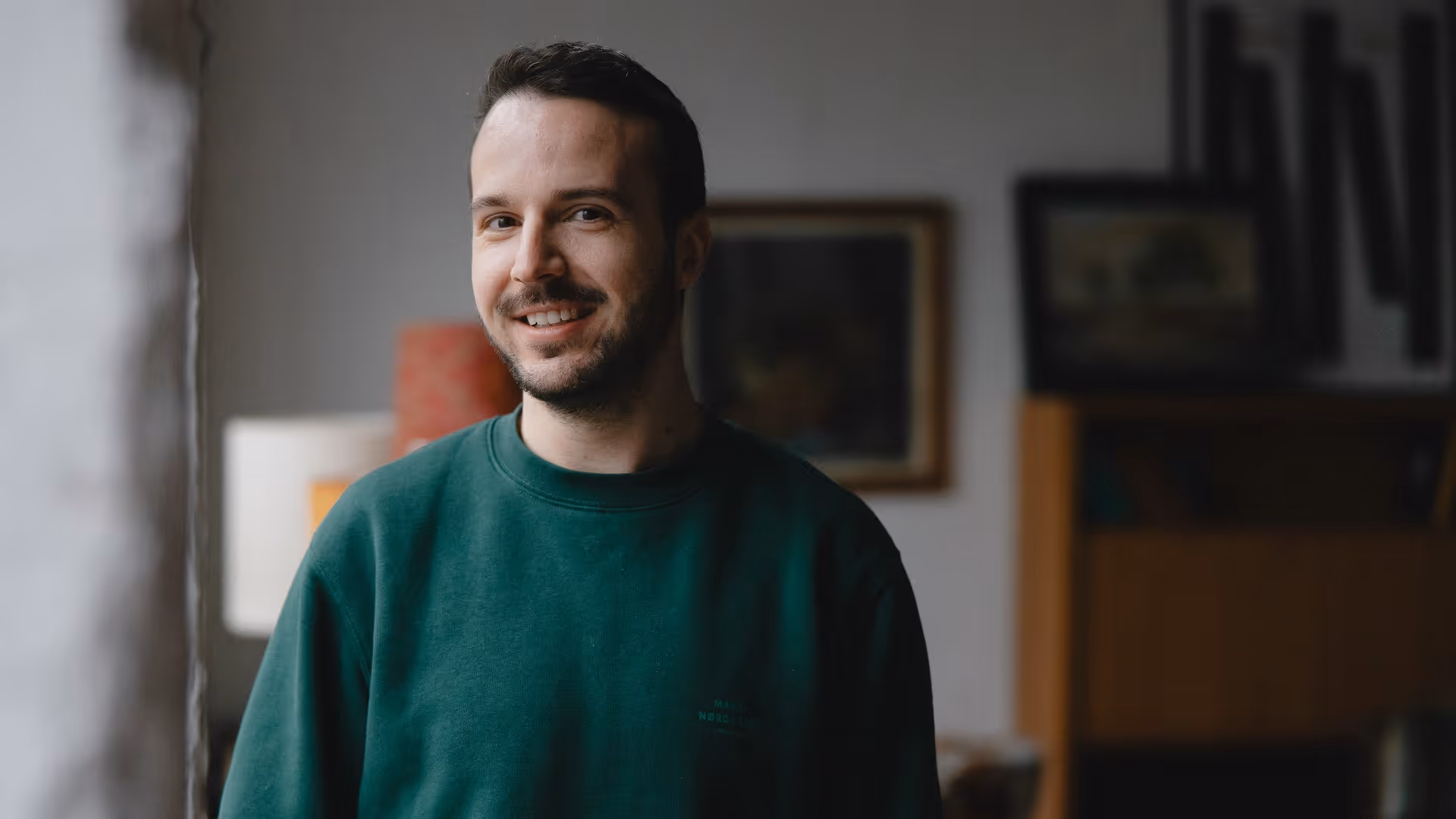 Ein lächelnder Mann mit kurzem dunklem Haar und Bart trägt einen grünen Pullover vor einem unscharfen Hintergrund mit Bildern und Regalen.